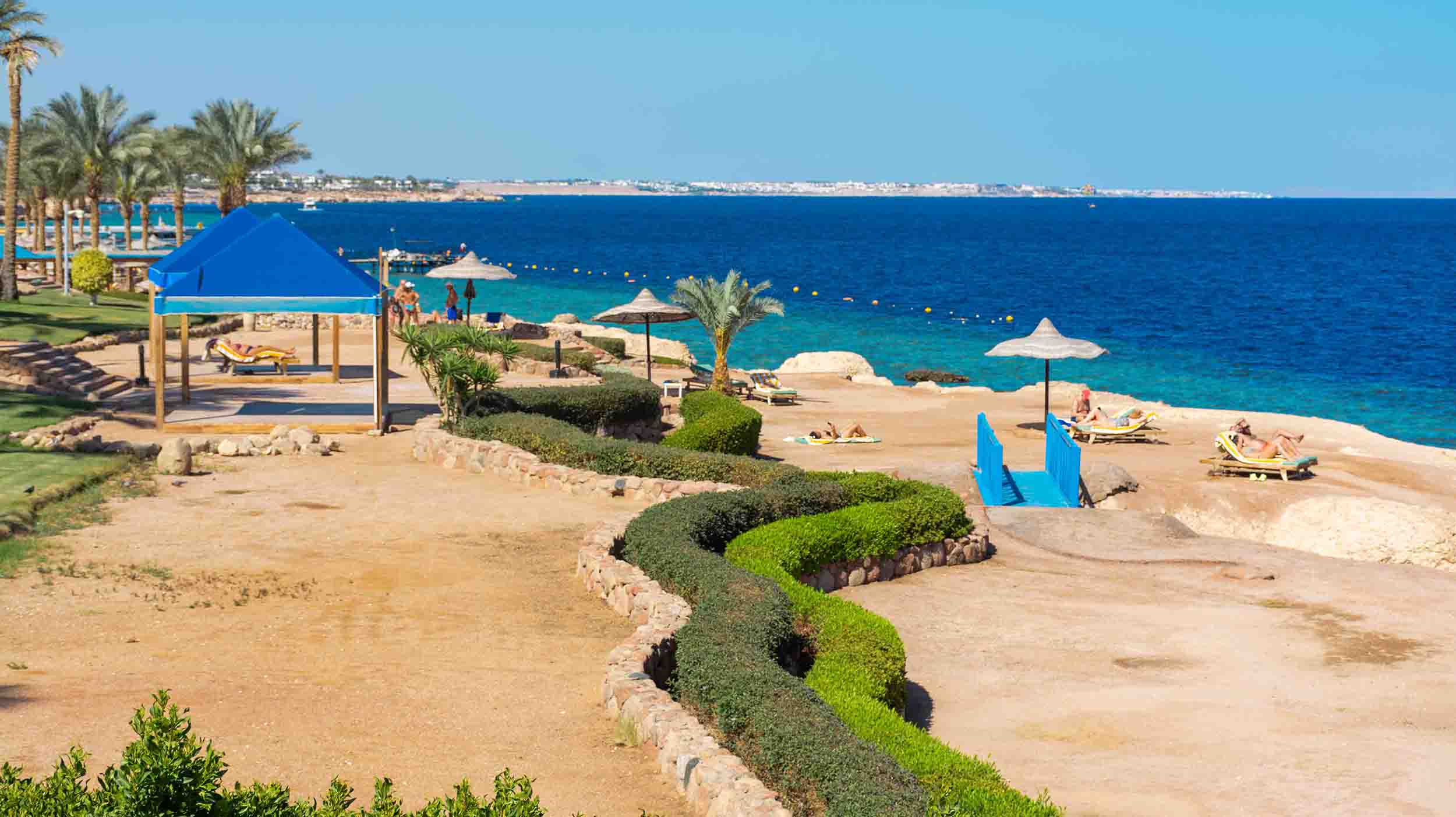 El contraste del Sinaí: Calma y descanso en Sharm el-Sheij Vista panorámica de la playa El Fanar en Sharm el-Sheij, con sombrillas de paja, camastros y una carpa azul sobre la costa arenosa, bordeada por un mar Rojo de aguas turquesas y palmeras
