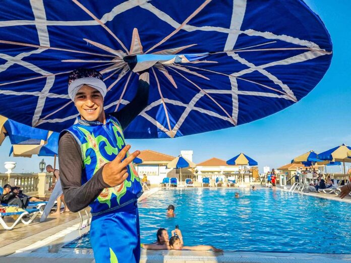 Un bailarín sonriente con vestimenta folclórica azul y turbante blanco realiza un espectáculo de Tanura sosteniendo una gran falda extendida con patrones geométricos, junto a una piscina de hotel en Sharm el-Sheij con huéspedes nadando al fondo