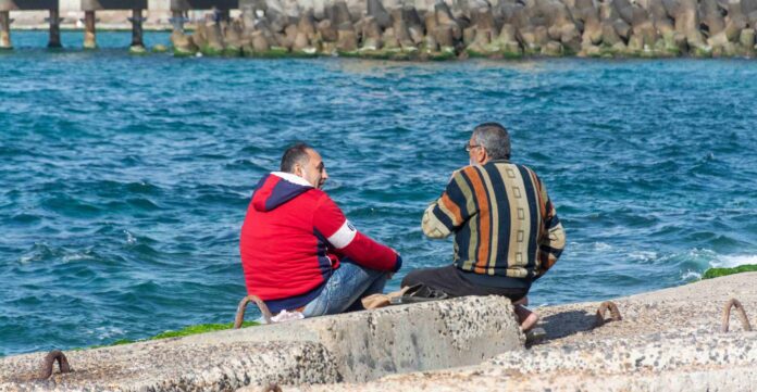 Dos hombres de mediana edad sentados de espaldas sobre un rompeolas de hormigón, cerca de la Fortaleza de Qaitbay en Alejandría, conversando tranquilamente mientras contemplan el oleaje del mar Mediterráneo. Uno viste una sudadera roja con capucha y el otro un suéter de rayas multicolores