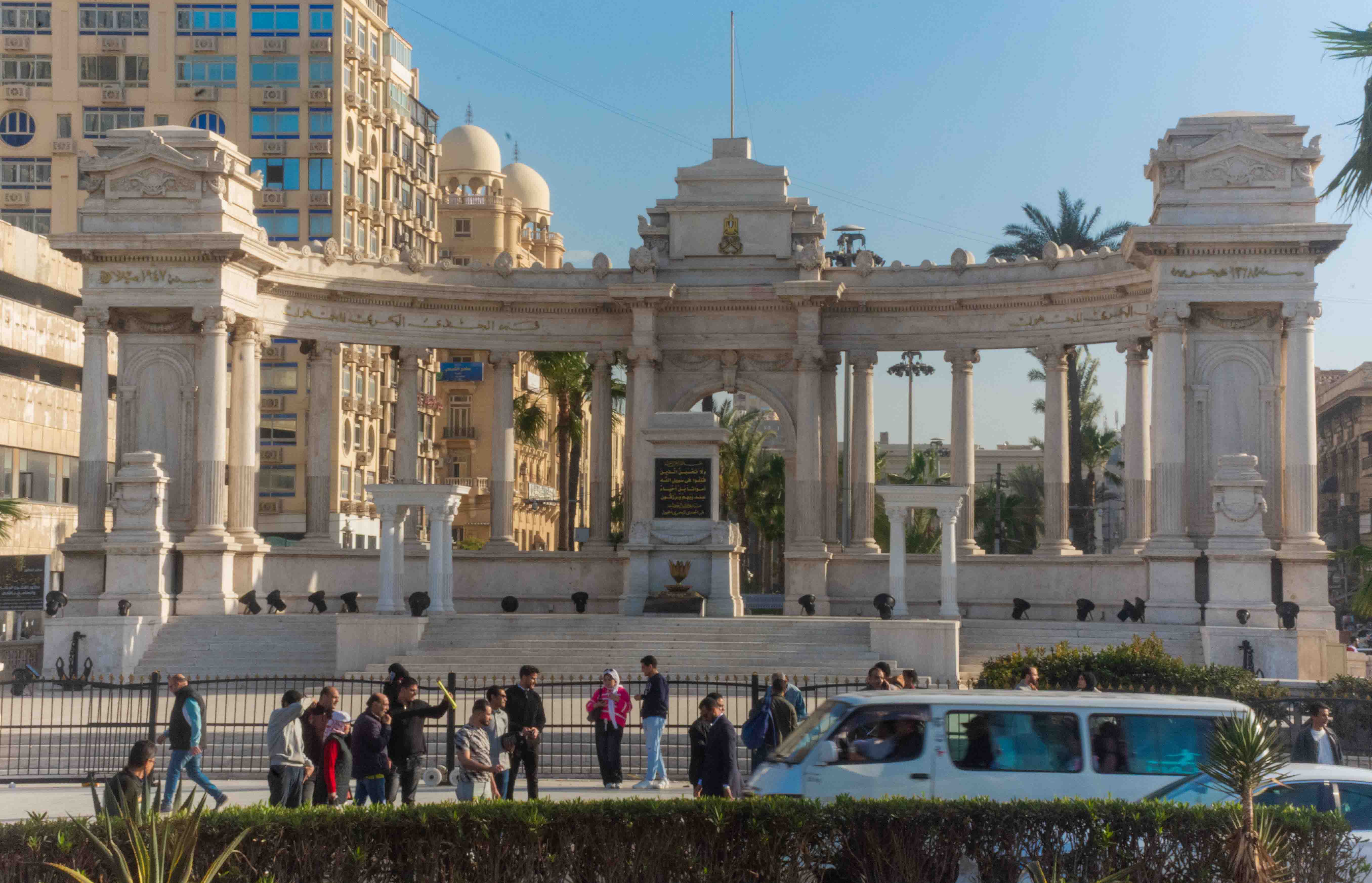 Monumento al Soldado Desconocido en Alejandría, compuesto por una gran columnata semicircular de mármol blanco de estilo neoclásico con un arco central que resguarda una lápida conmemorativa, mientras peatones caminan por la plaza frontal y un microbús blanco transita por la calle