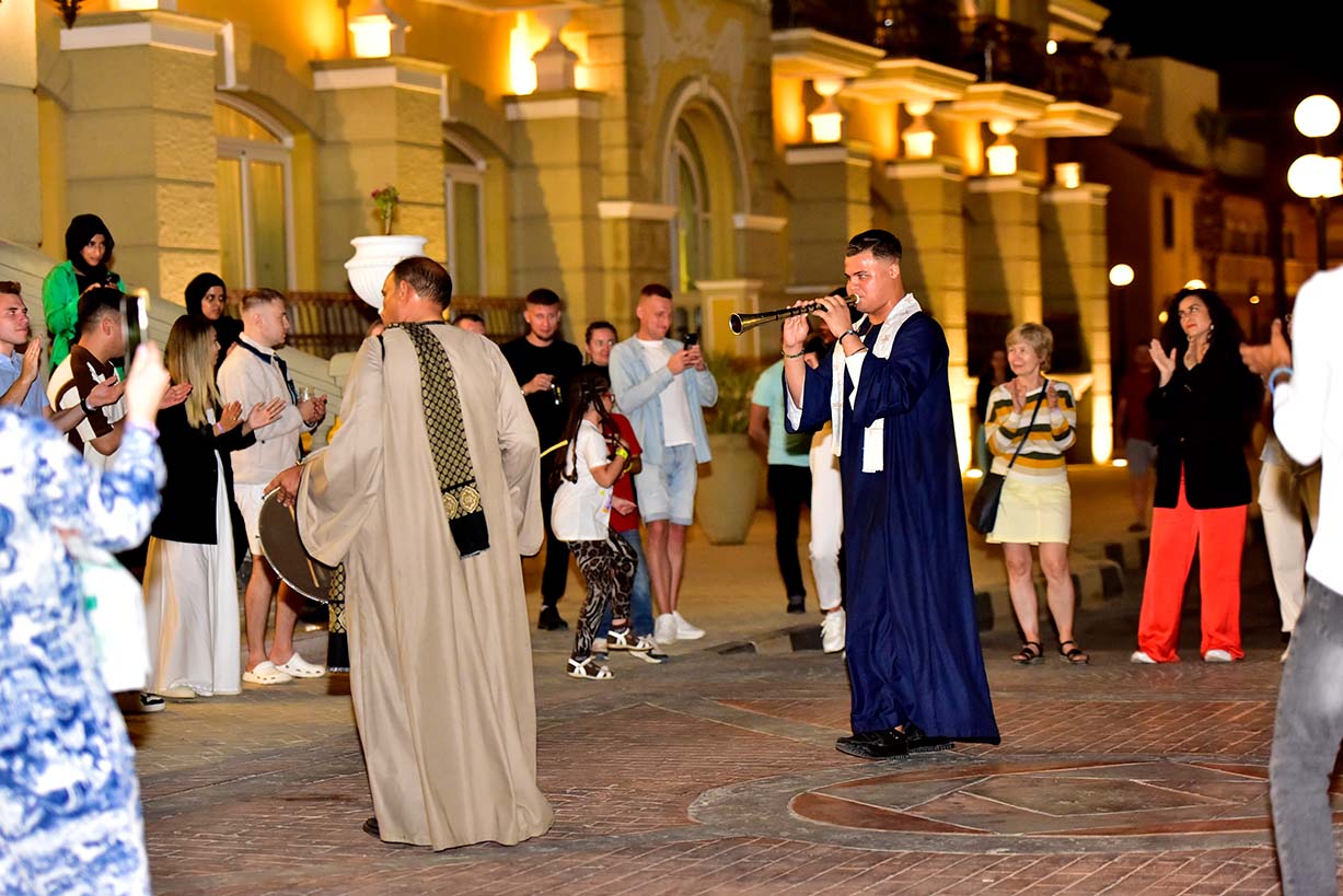 Músicos egipcios vestidos con túnicas tradicionales tocan instrumentos de viento y percusión por la noche frente a un grupo de turistas que aplauden en una calle iluminada de Sharm el-Sheij