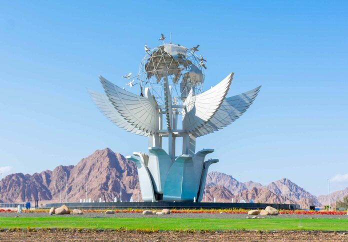 Un gran monumento blanco y moderno con forma de flor de loto gigante de cuyas hojas brotan unas alas extendidas que sostienen un globo terráqueo metálico rodeado de palomas de la paz en vuelo, situado en una plaza verde con las montañas áridas del Sinaí bajo un cielo azul despejado al fondo