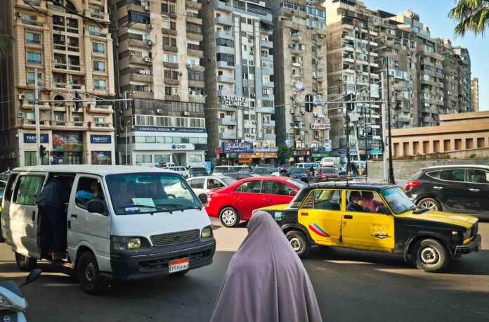 Tráfico denso en la plaza Víctor Manuel en Smouha, Alejandría, mostrando una furgoneta blanca, un taxi clásico negro y amarillo, y edificios residenciales altos al fondo