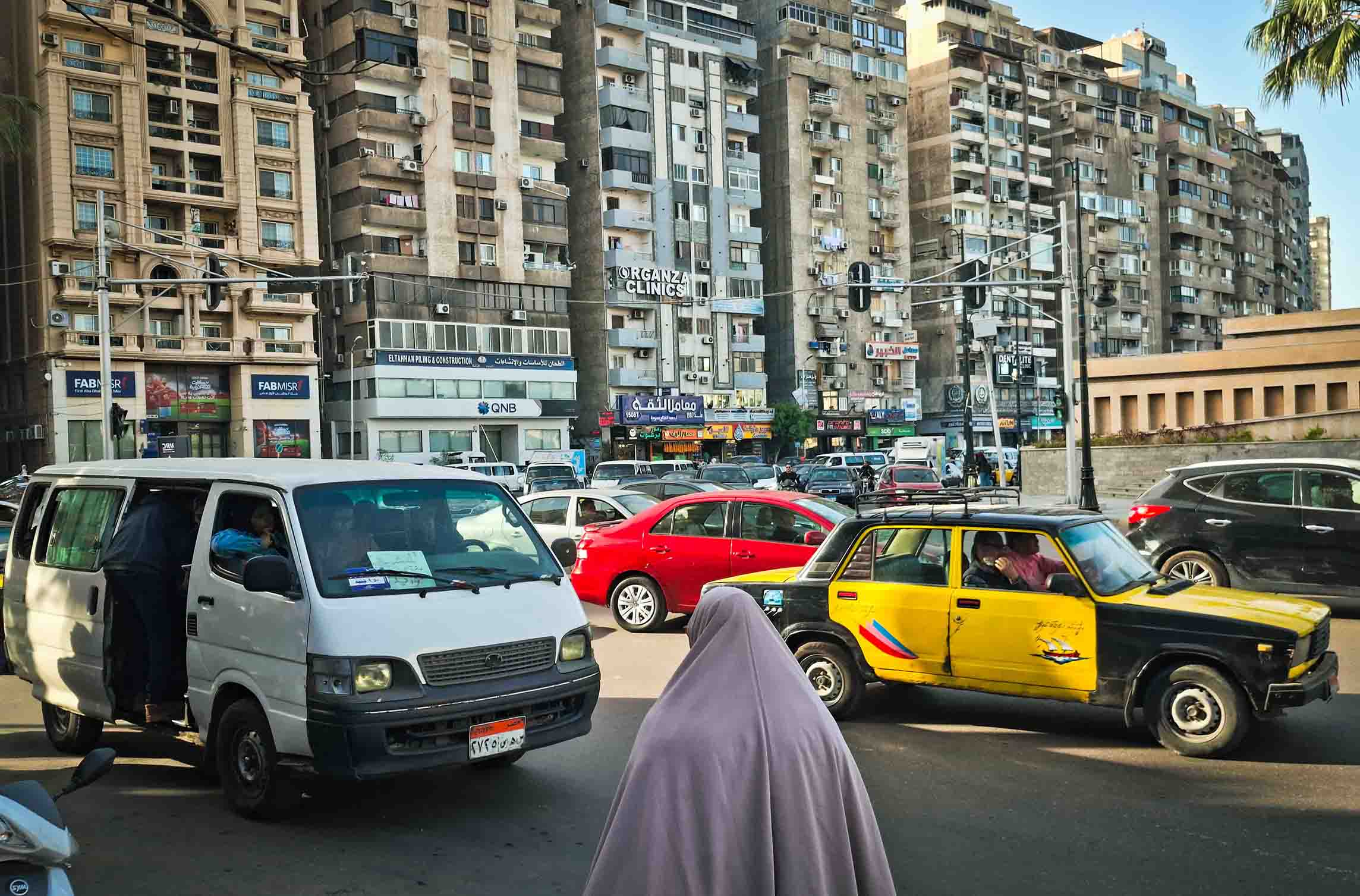 Tráfico denso en la plaza Víctor Manuel en Smouha, Alejandría, mostrando una furgoneta blanca, un taxi clásico negro y amarillo, y edificios residenciales altos al fondo