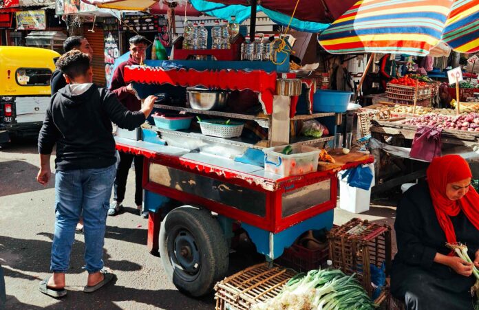 Un colorido carrito callejero de comida decorado en rojo y azul en un mercado concurrido; un joven de espaldas espera su comida mientras una mujer sentada a un costado limpia cebollas verdes frescas