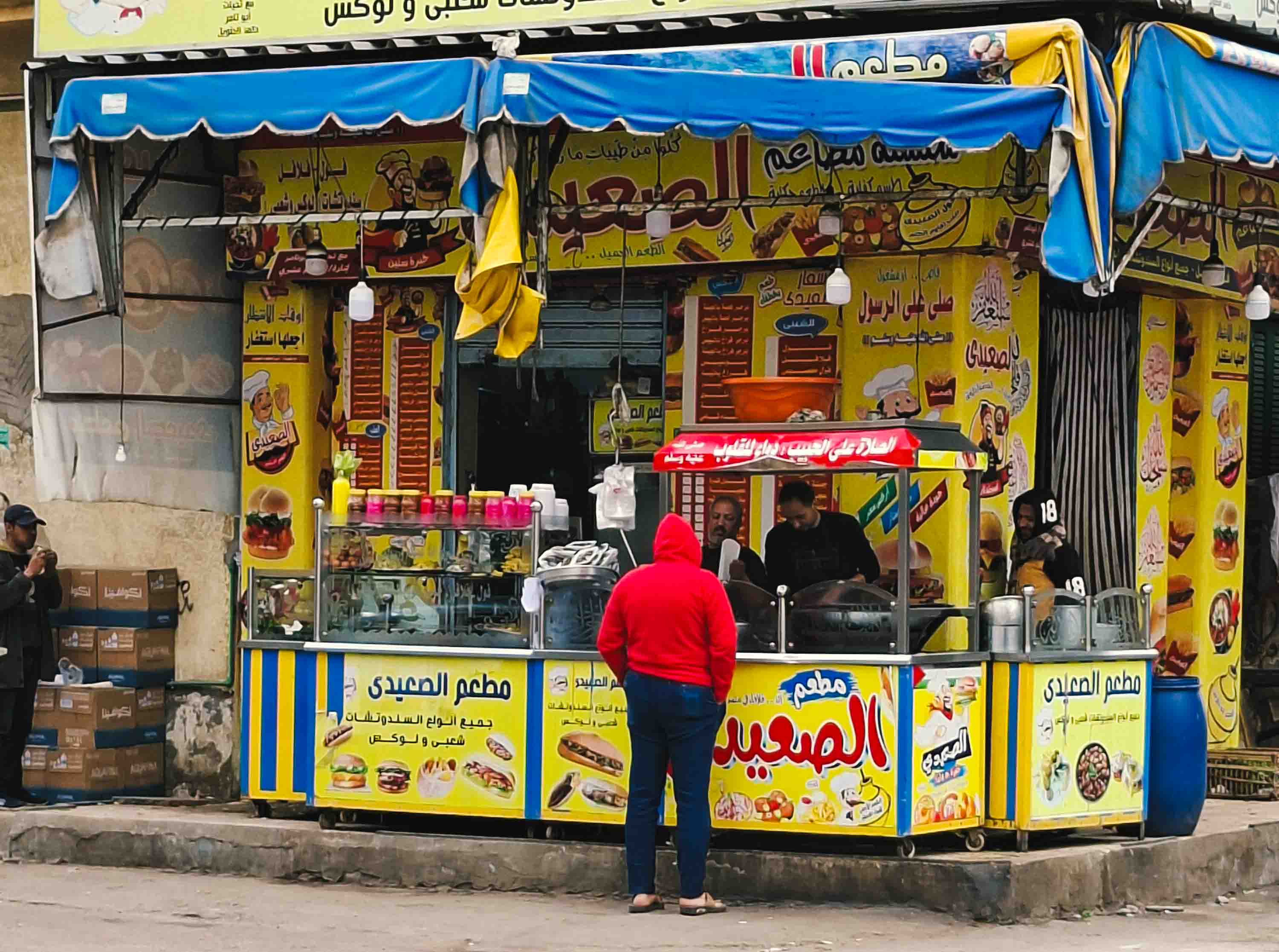 Fachada de un local de comida rápida callejera de color amarillo brillante con letreros en árabe; un cliente de espaldas con sudadera roja espera frente al mostrador donde los cocineros preparan los alimentos