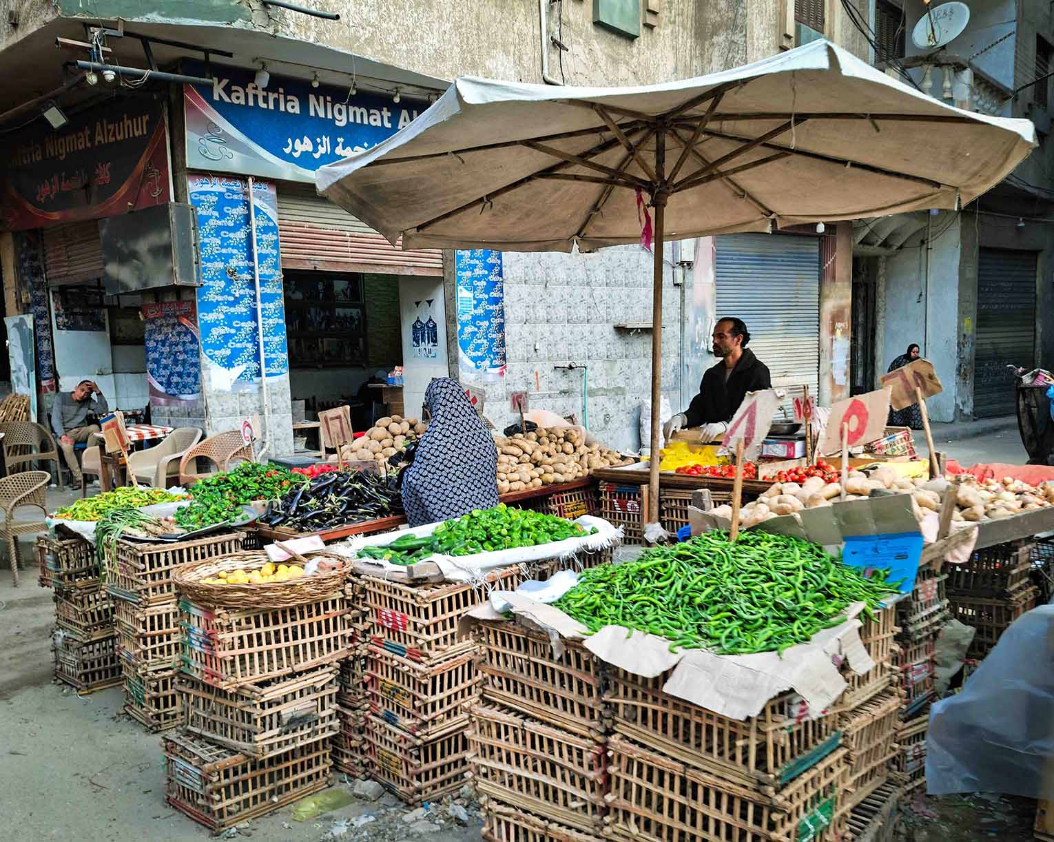 Un puesto callejero en Alejandría cubierto por una gran sombrilla blanca expone una gran variedad de verduras frescas como pimientos verdes, berenjenas, papas y tomates, organizados en cajas de madera apiladas frente a una cafetería local