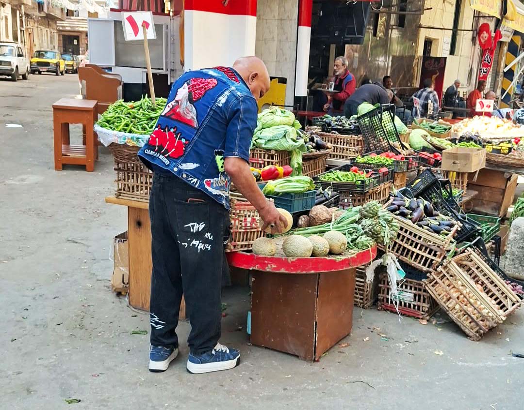 Un vendedor ambulante de espaldas, vestido con pantalones negros y un chaleco de mezclilla decorado, acomoda melones en una mesa redonda dentro de un mercado callejero en Alejandría, rodeado de cajas de madera llenas de verduras como pimientos, alcachofas y berenjenas