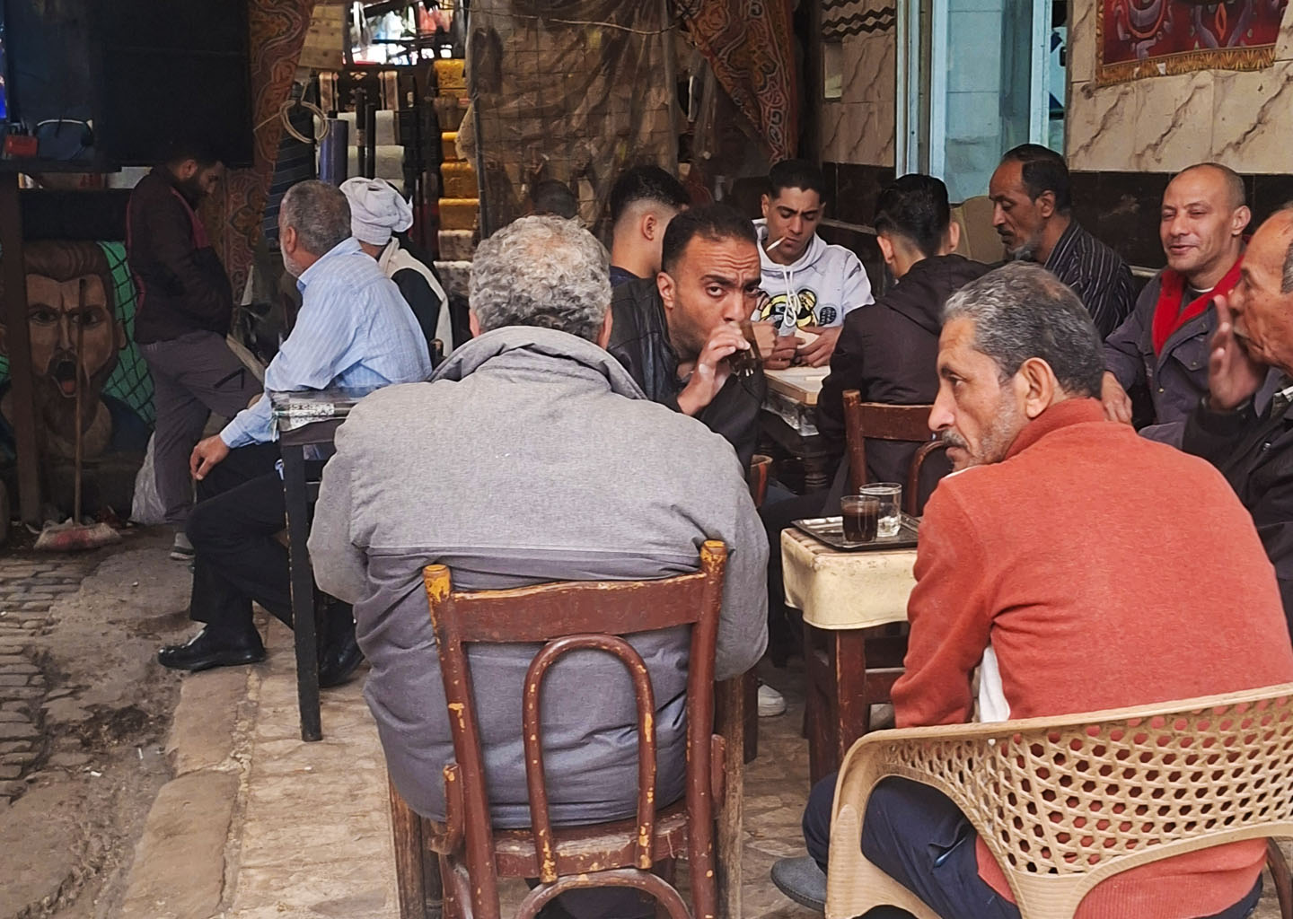 Un grupo de hombres egipcios de distintas edades sentados muy juntos en sillas de madera y plástico dentro de un café tradicional en Alejandría, conversando y bebiendo pequeños vasos de té servidos en mesas rústicas