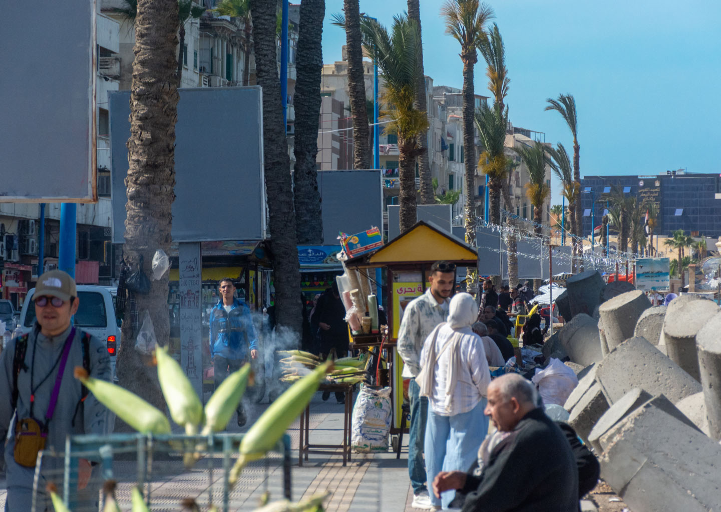Personas paseando por la Cornisa de Alejandría con palmeras, puestos callejeros y bloques de hormigón junto al mar