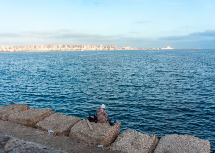 Un hombre con sombrero y camisa de cuadros pesca sentado sobre grandes bloques de hormigón en la costa de Alejandría, con el perfil urbano de la ciudad al fondo bajo un cielo despejado