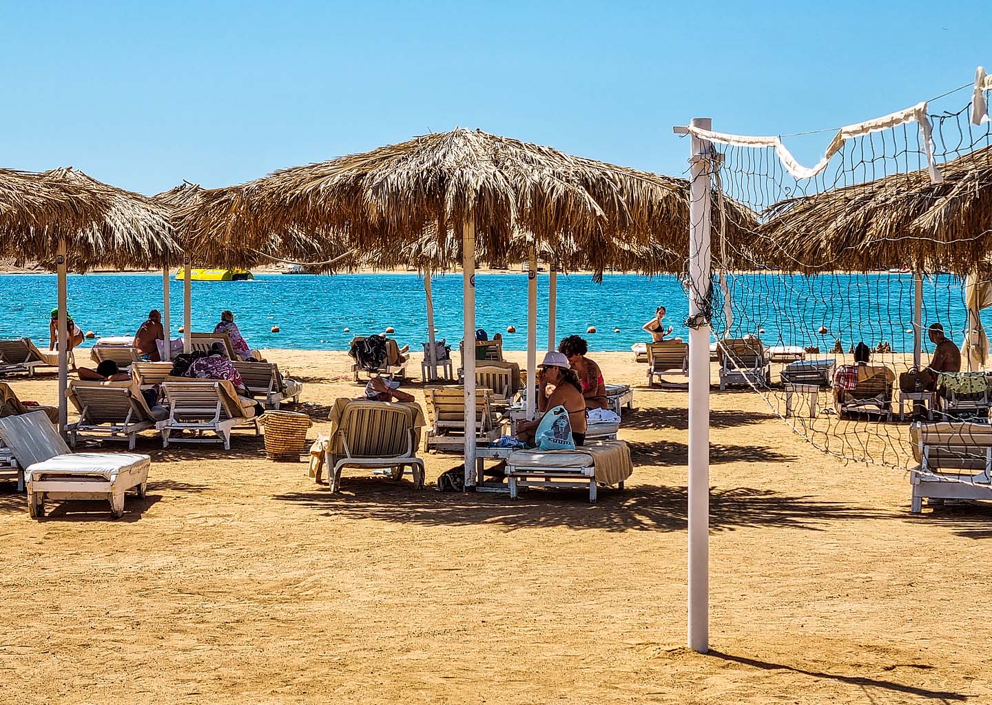 La playa Bella Vita turística en Sharm el-Sheij con sombrillas de paja, tumbonas ocupadas por bañistas, una red de voleibol de playa en primer plano y aguas de azul turquesa al fondo