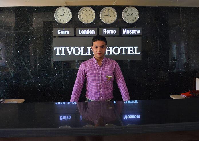 Un joven recepcionista egipcio posa tras el mostrador del Tivoli Hotel en Sharm el-Sheij. En el fondo, se aprecian relojes de pared que muestran la hora de El Cairo, Londres, Roma y Moscú, reflejando la naturaleza internacional del sector turístico en Egipto