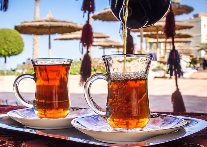 Primer plano de una tetera oscura vertiendo té caliente y ambarino en uno de los dos pequeños vasos de cristal con asa colocados sobre platillos decorados en una bandeja metálica, con un fondo difuminado de sombrillas de playa de paja bajo un cielo despejado