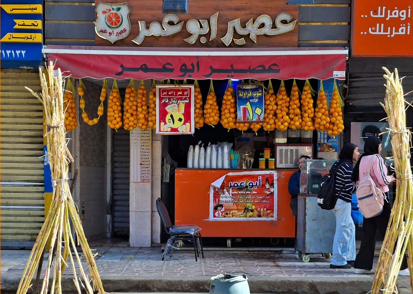 Fachada de una tienda local de zumos en Egipto llamada "Abo Omar", con bolsas de naranjas colgadas del toldo y cañas de azúcar atadas en la entrada, mientras dos jóvenes caminan por delante
