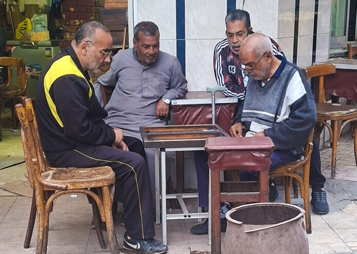 Cuatro hombres mayores reunidos en la terraza de un café local egipcio alrededor de una mesa de madera, concentrados mientras juegan una partida de backgammon tradicional