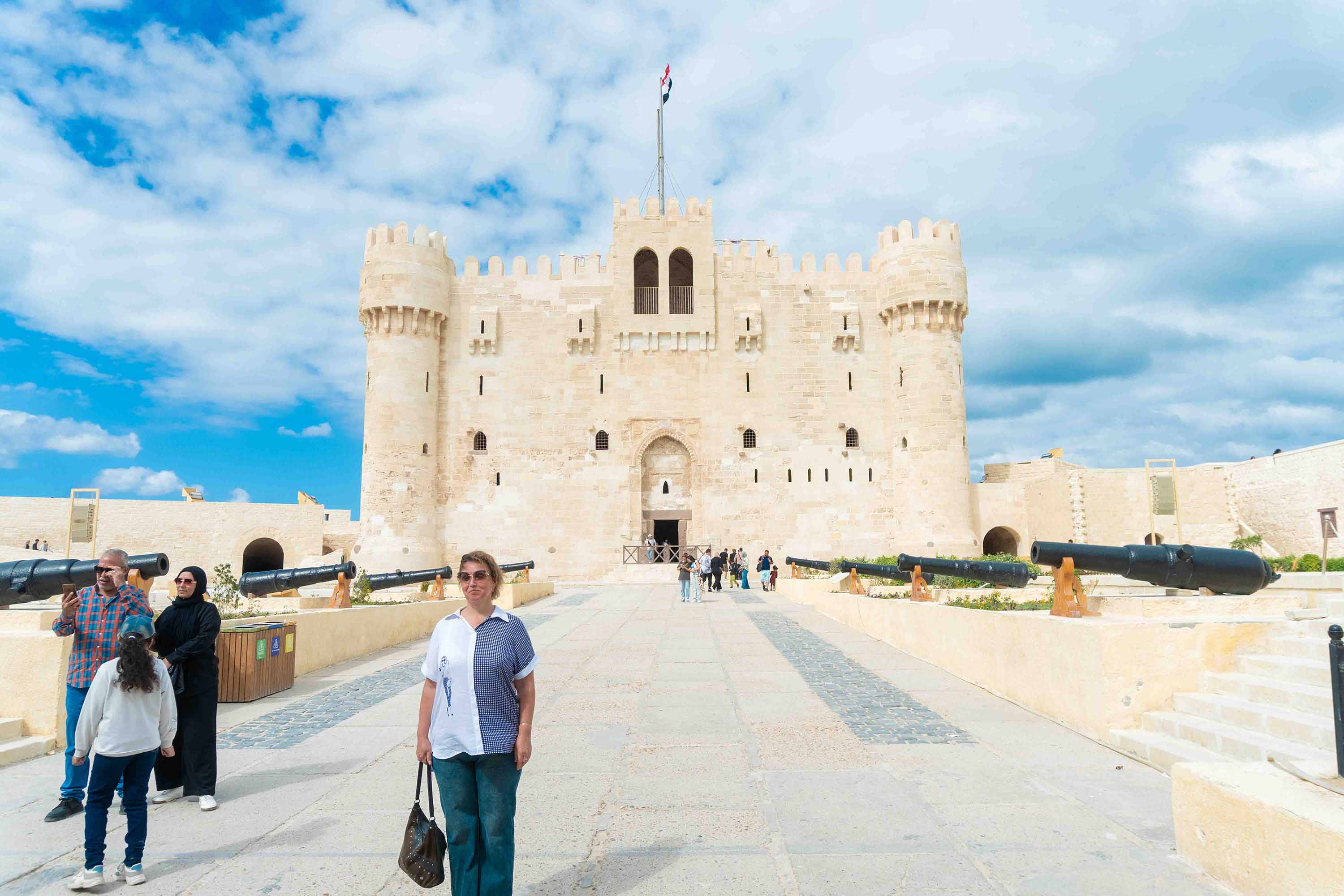 Una turista posa sonriente frente a la imponente fachada de piedra de la Ciudadela de Qaitbay en Alejandría, rodeada de antiguos cañones y otros visitantes bajo un cielo parcialmente nublado