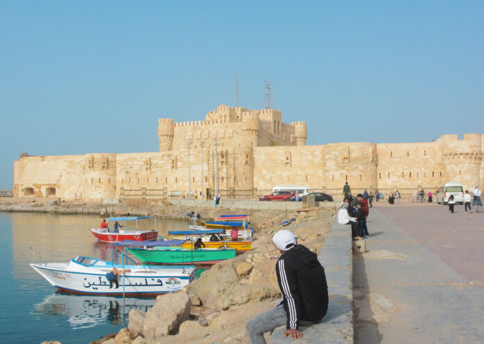Vista de la Ciudadela de Qaitbay en Alejandría durante el verano en Egipto, con barcas de colores amarradas en el agua y personas paseando junto a la fortaleza bajo un cielo despejado